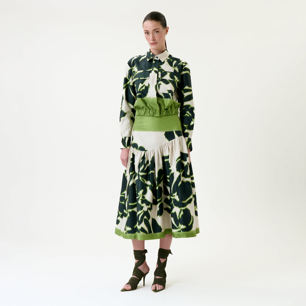 A woman stands against a white background, wearing the Libby Skirt—a long-sleeved shirt and matching skirt set featuring a green and dark abstract floral pattern, complete with a green waistband—and pairing it with green heeled sandals.