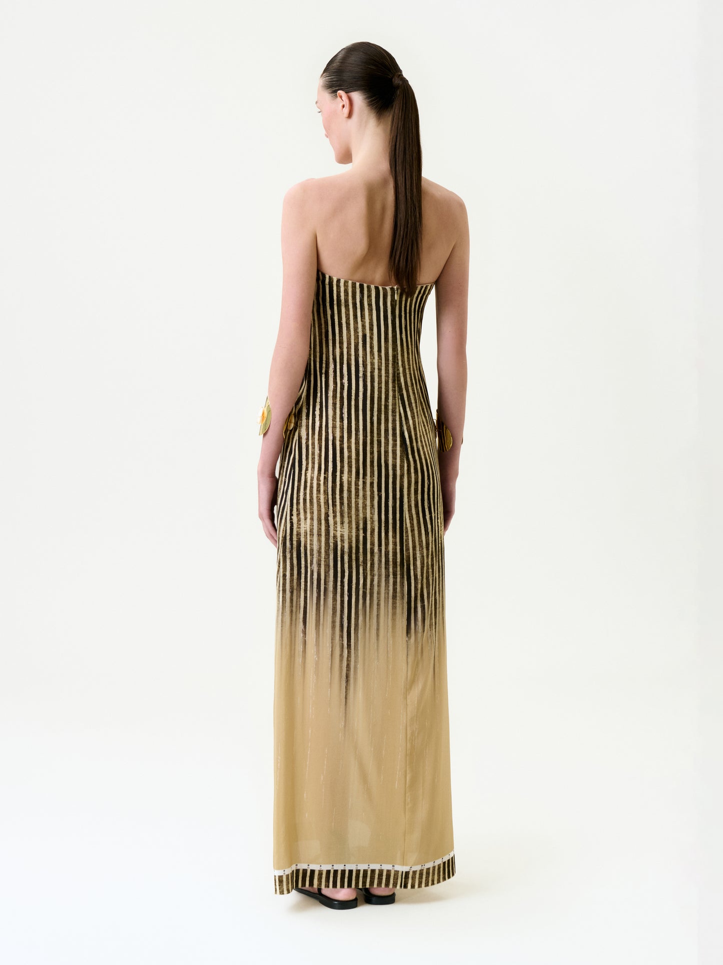 A person with long brown hair in a low ponytail faces away from the camera, wearing the Lirit Dress—a strapless, floor-length maxi crafted from Italian fabric with vertical black stripes on a beige background—against a plain white backdrop.