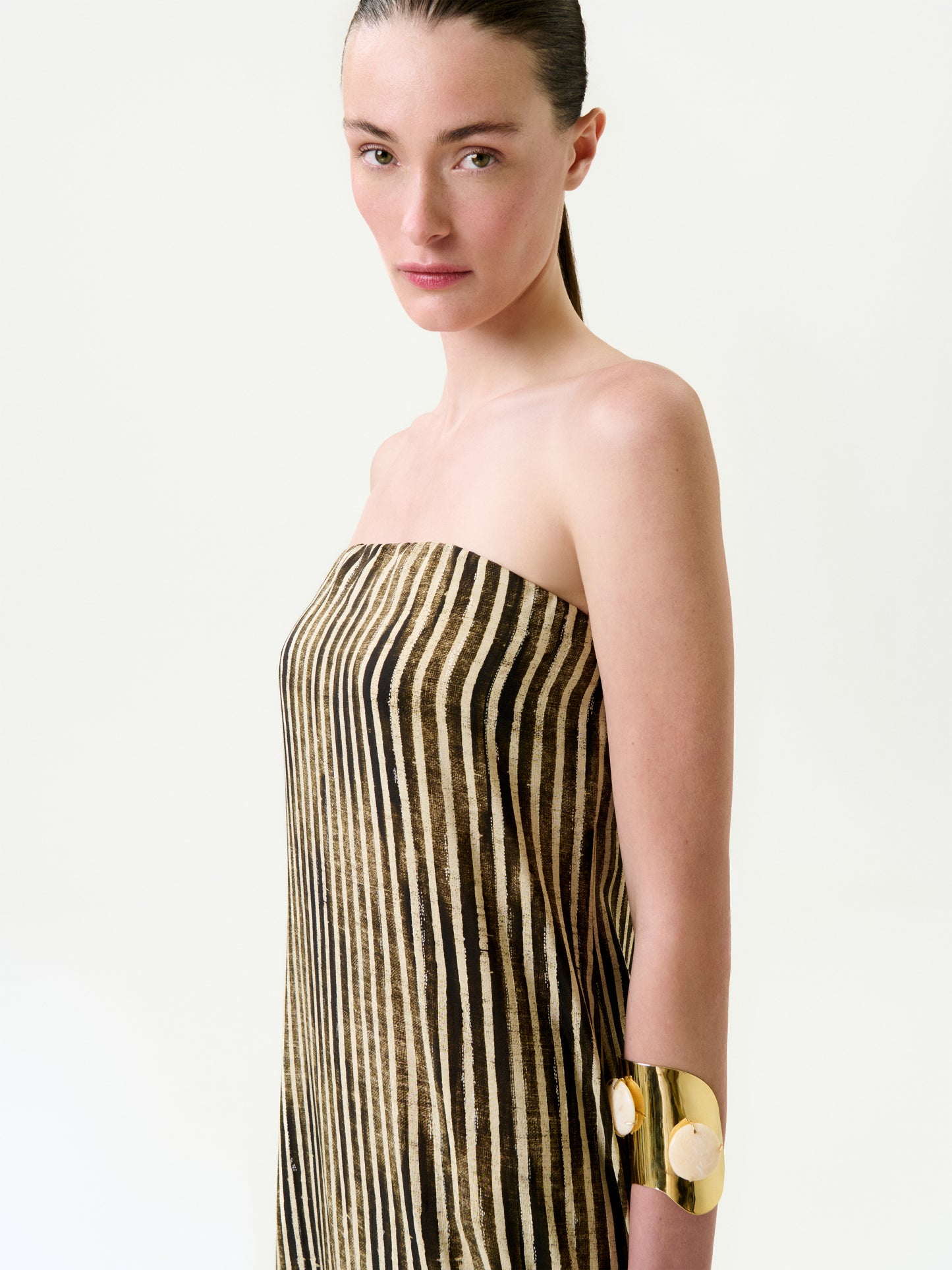 A woman with straight brown hair wears the Lirit Dress, a strapless maxi made from luxurious Italian fabric with vertical black and gold stripes, paired with a bold gold cuff bracelet against a plain light background.