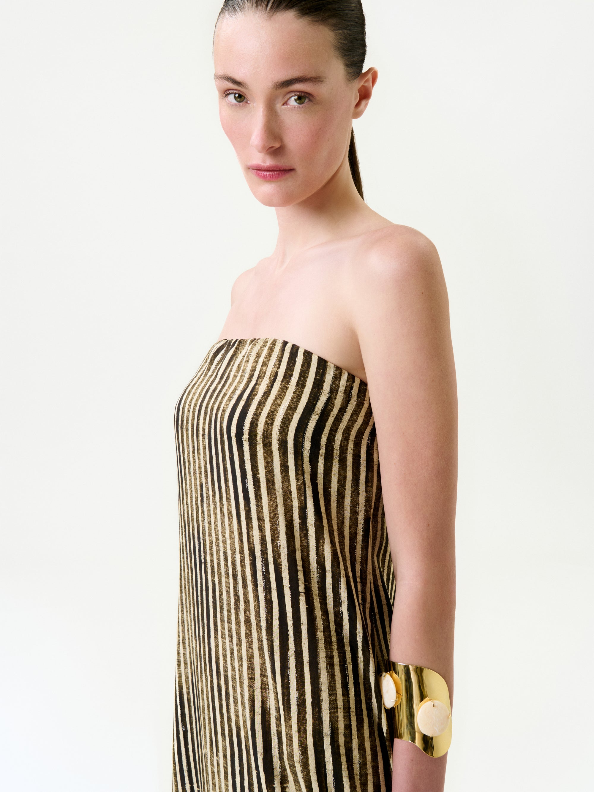 A woman with straight brown hair wears the Lirit Dress, a strapless maxi made from luxurious Italian fabric with vertical black and gold stripes, paired with a bold gold cuff bracelet against a plain light background.