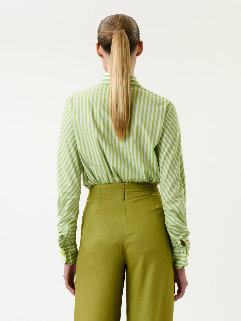 A person with long blonde hair in a ponytail, wearing the Lucelly Shirt tucked into high-waisted olive green pants, is shown from the back against a plain white background.