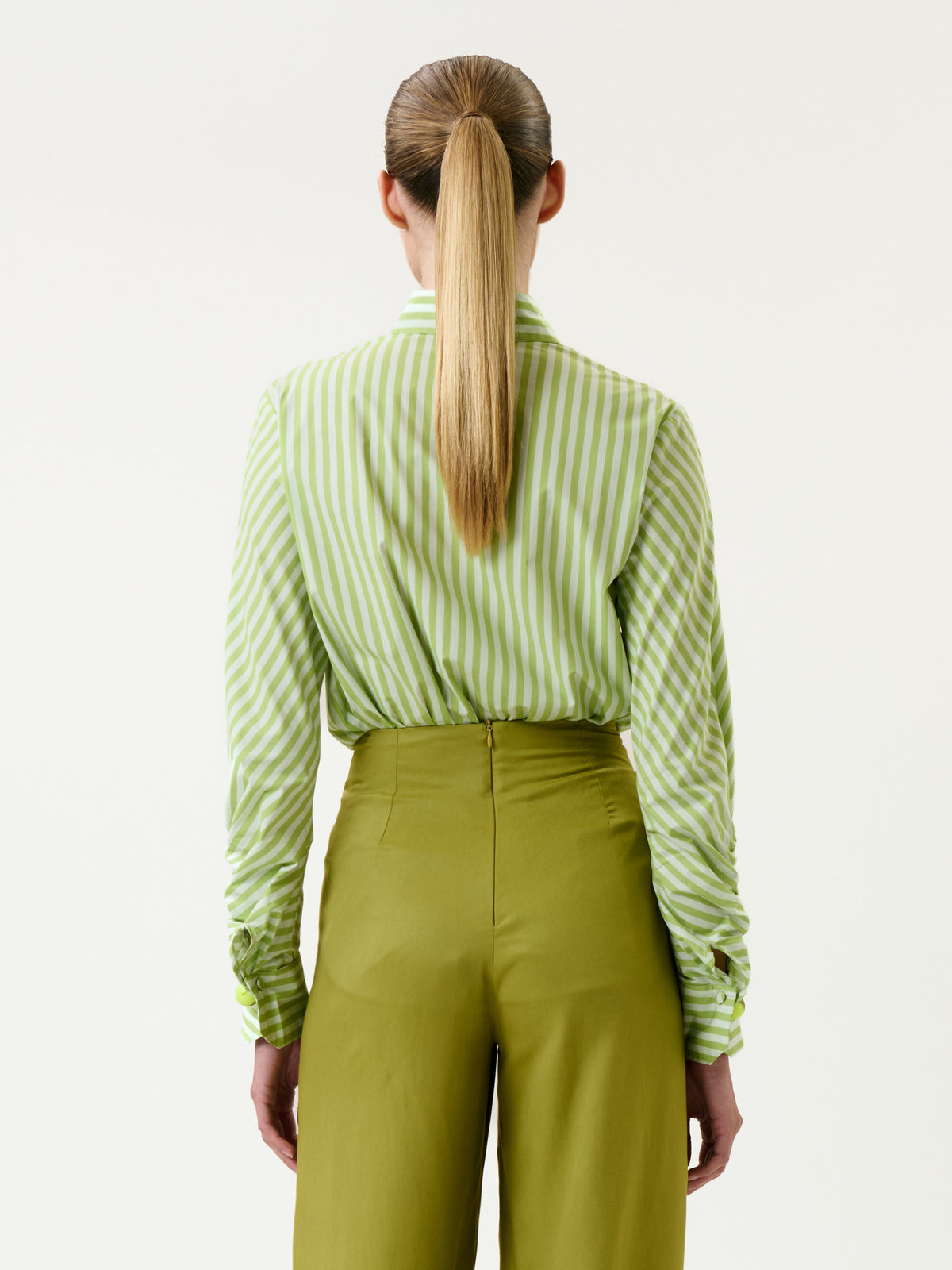 A person with long blonde hair in a ponytail, wearing the Lucelly Shirt tucked into high-waisted olive green pants, is shown from the back against a plain white background.