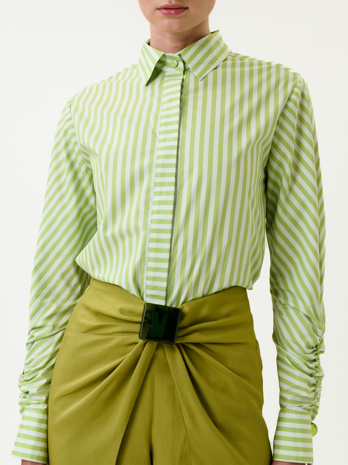 A person wearing the Lucelly Shirt—a light green and white vertically striped button-up—tucked into high-waisted, olive green pants with a draped front and a black rectangular belt buckle.