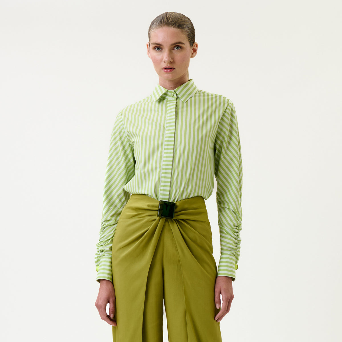 A woman stands facing forward, wearing the Lucelly Shirt—a green-and-white striped top—tucked into high-waisted, olive-green trousers featuring a front knot and wide belt. She has slicked-back hair and a neutral expression against a plain background.