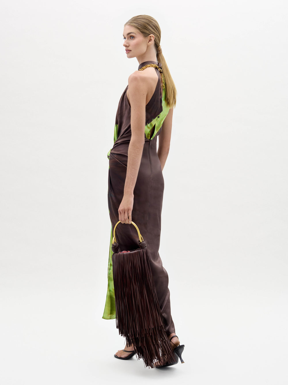 A woman in the Tizziana Dress, a brown and green sleeveless style, holds a large fringed bag and wears black heeled sandals against a plain white background.