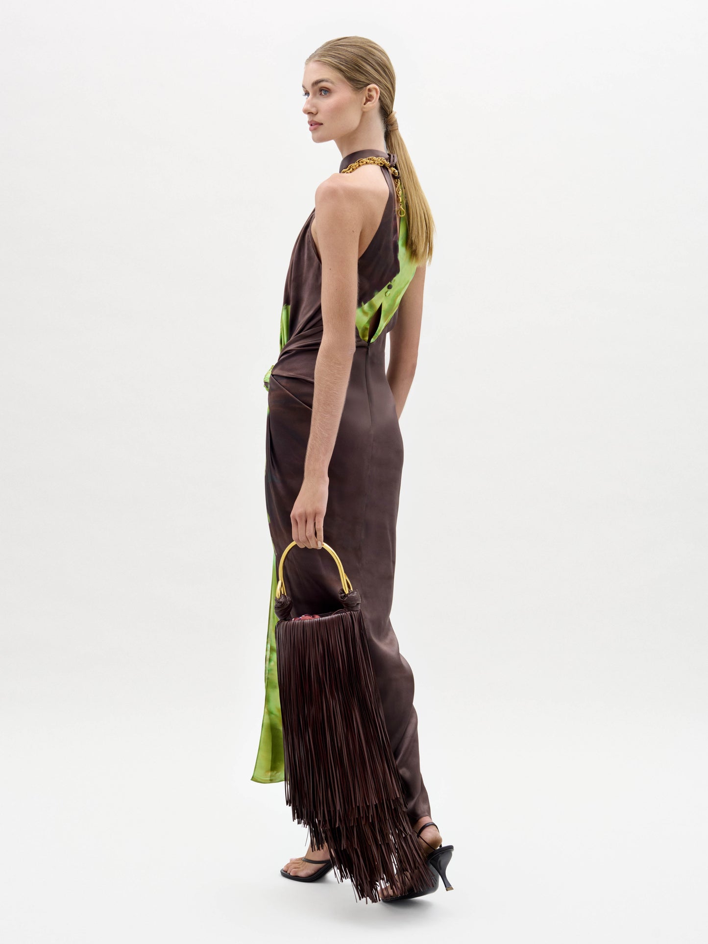 A woman in the Tizziana Dress, a brown and green sleeveless style, holds a large fringed bag and wears black heeled sandals against a plain white background.
