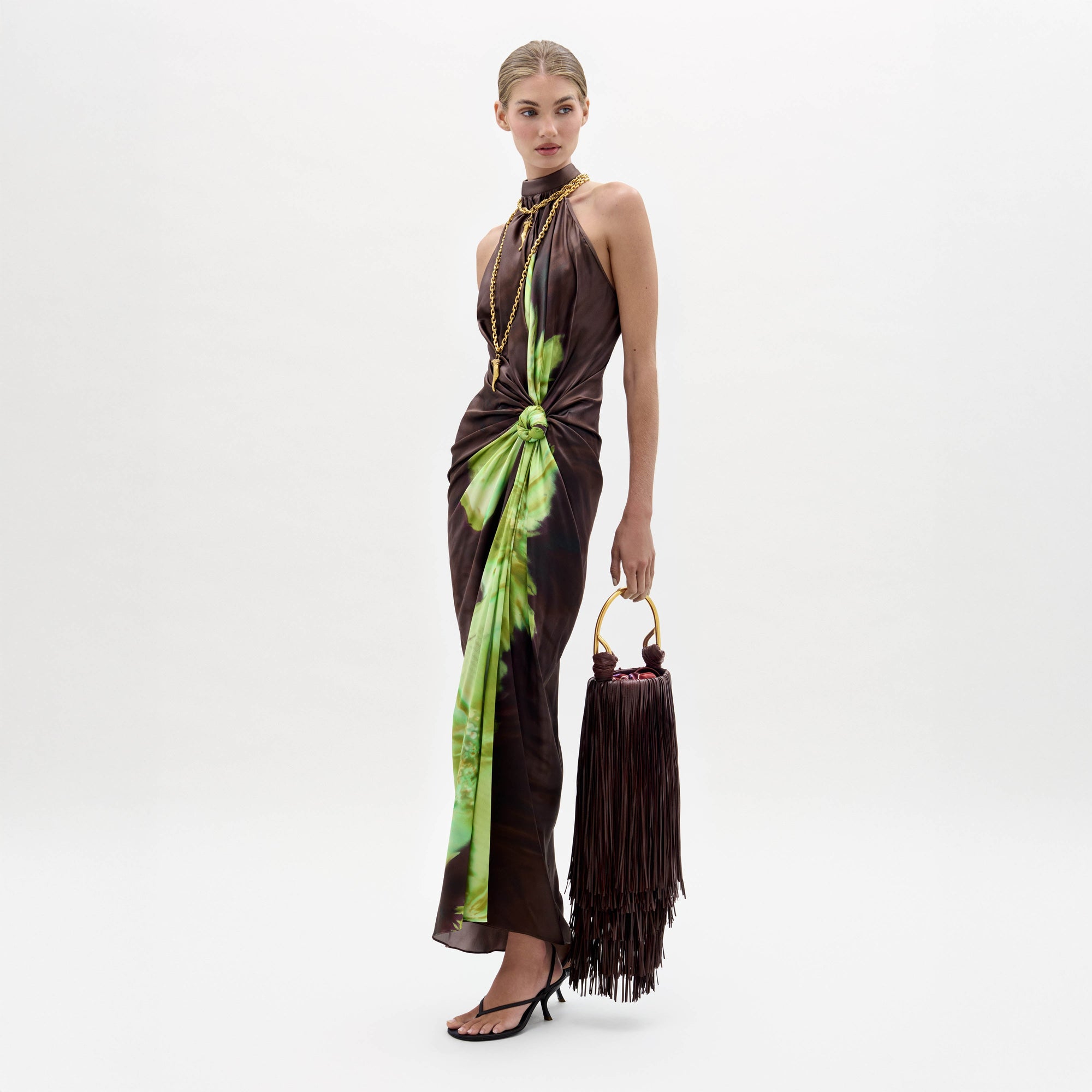 A woman in the Tizziana Dress, a brown and green sleeveless style, holds a large fringed bag and wears black heeled sandals against a plain white background.