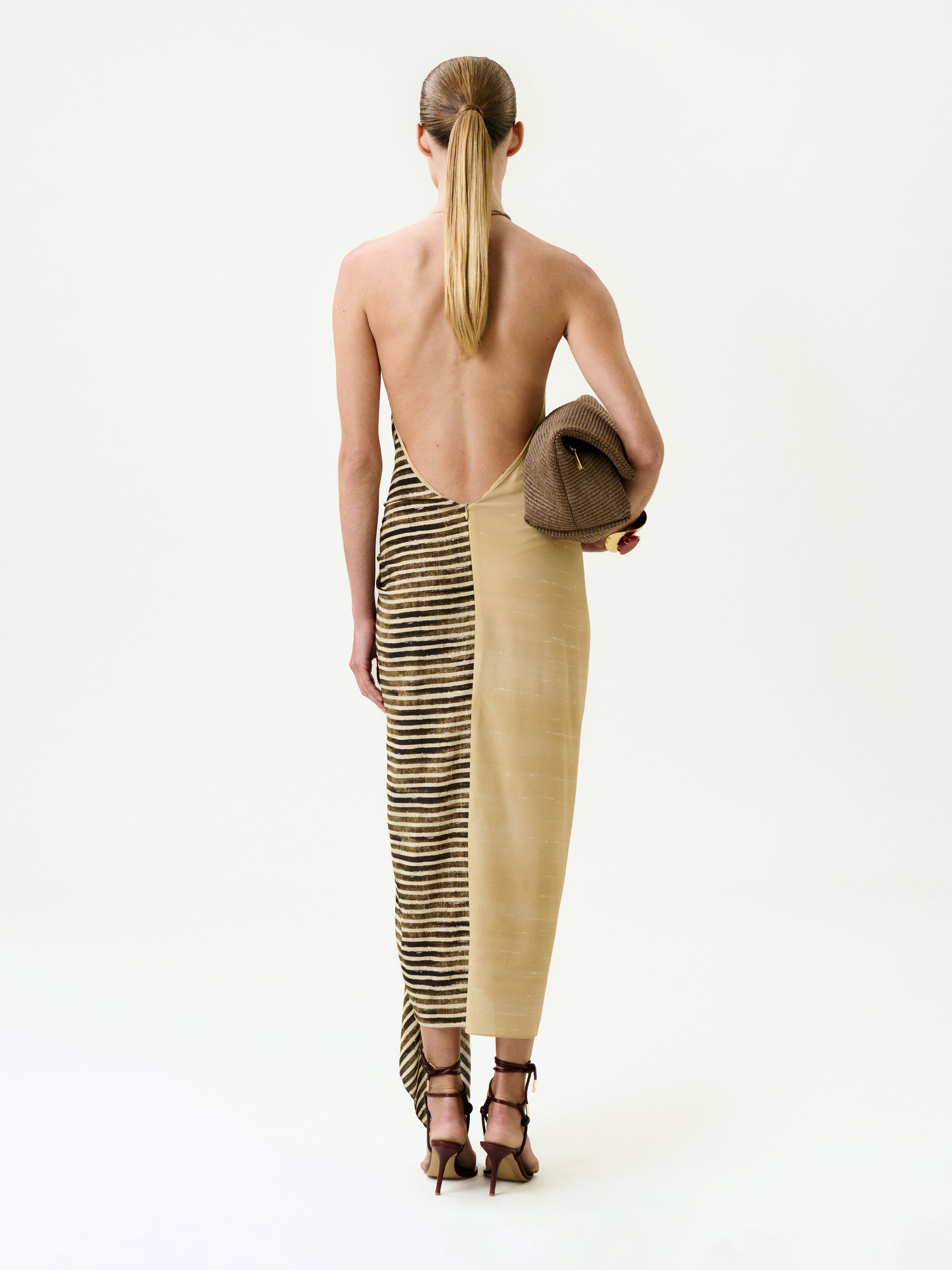 A person with slicked-back hair stands facing away, wearing the Lorde Dress—a backless, two-toned silk midi dress in beige and stripes—paired with a textured brown clutch and strappy high-heeled sandals.