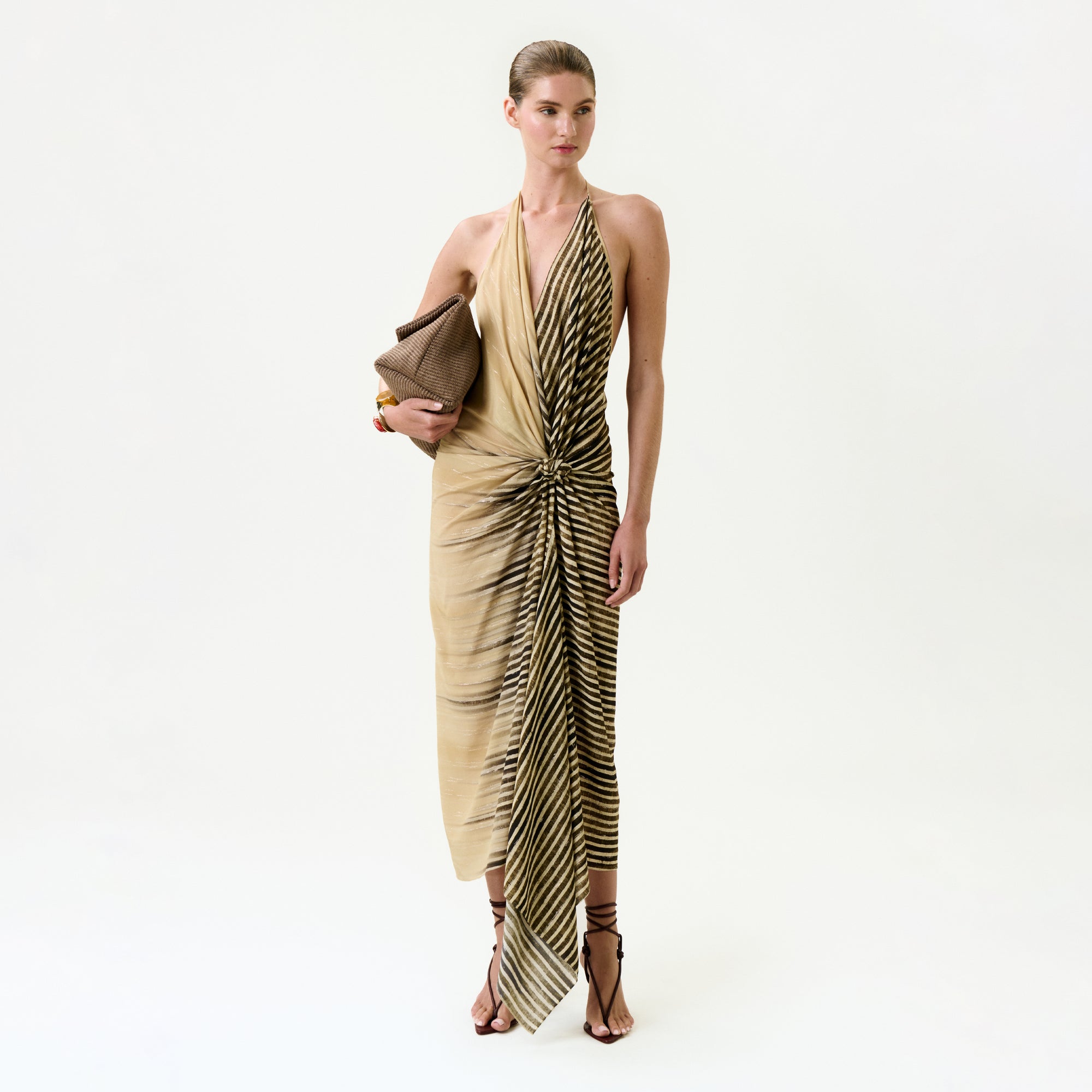 A woman stands against a plain background, wearing the Lorde Dress—a beige and black striped silk midi made from Italian fabric. She pairs it with strappy heeled sandals, gold bracelets, slicked-back hair, and a brown clutch.