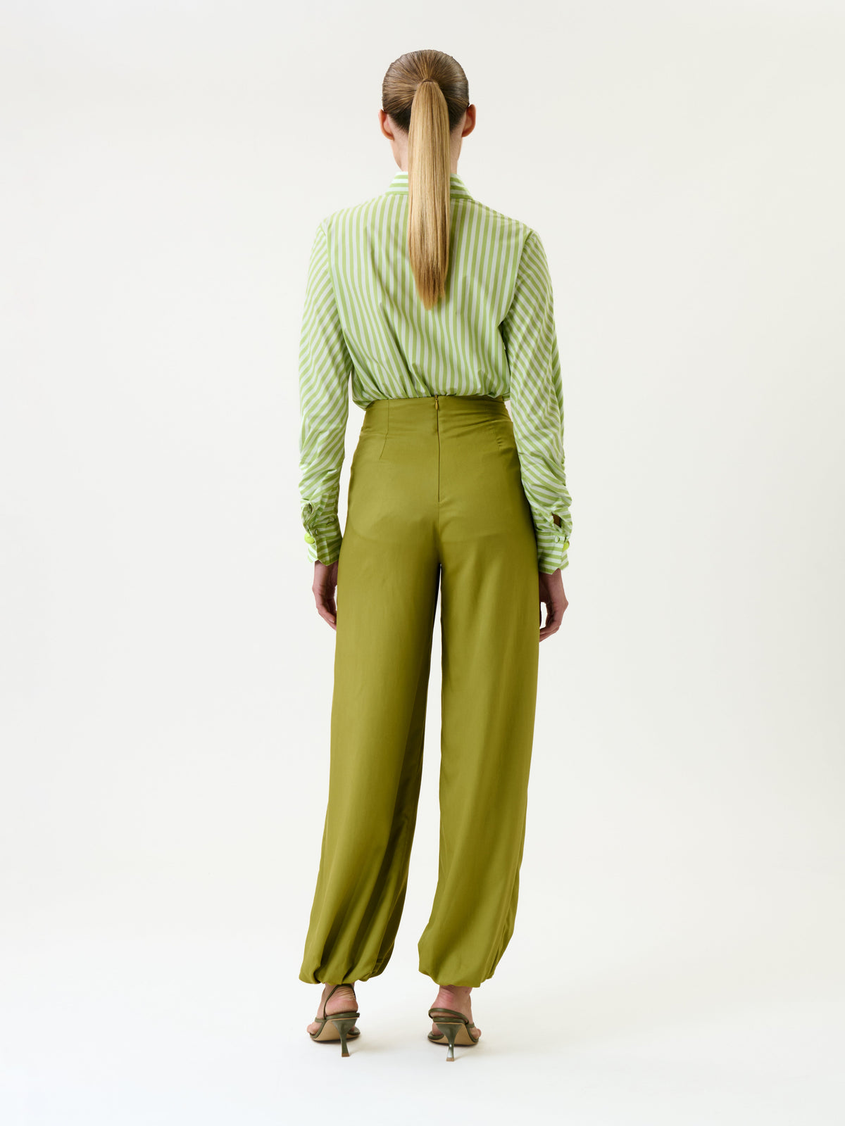 A woman stands with her back to the camera, wearing the Raimondo Pant—high-waisted, wide-leg olive trousers—paired with a green and white striped shirt and heeled sandals. Her hair is styled in a low ponytail, and she is set against a plain white background.