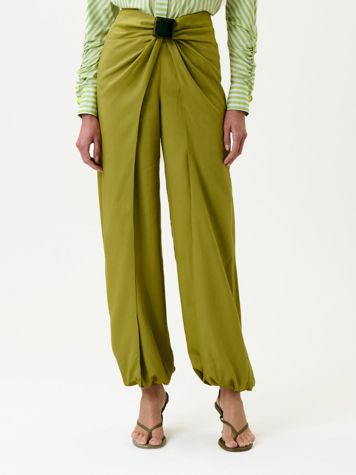 A person is shown from the waist down, wearing the Raimondo Pant in olive green with gathered details and a front buckle, paired with a light green and white striped shirt and open-toe sandals. They are standing against a plain white background.