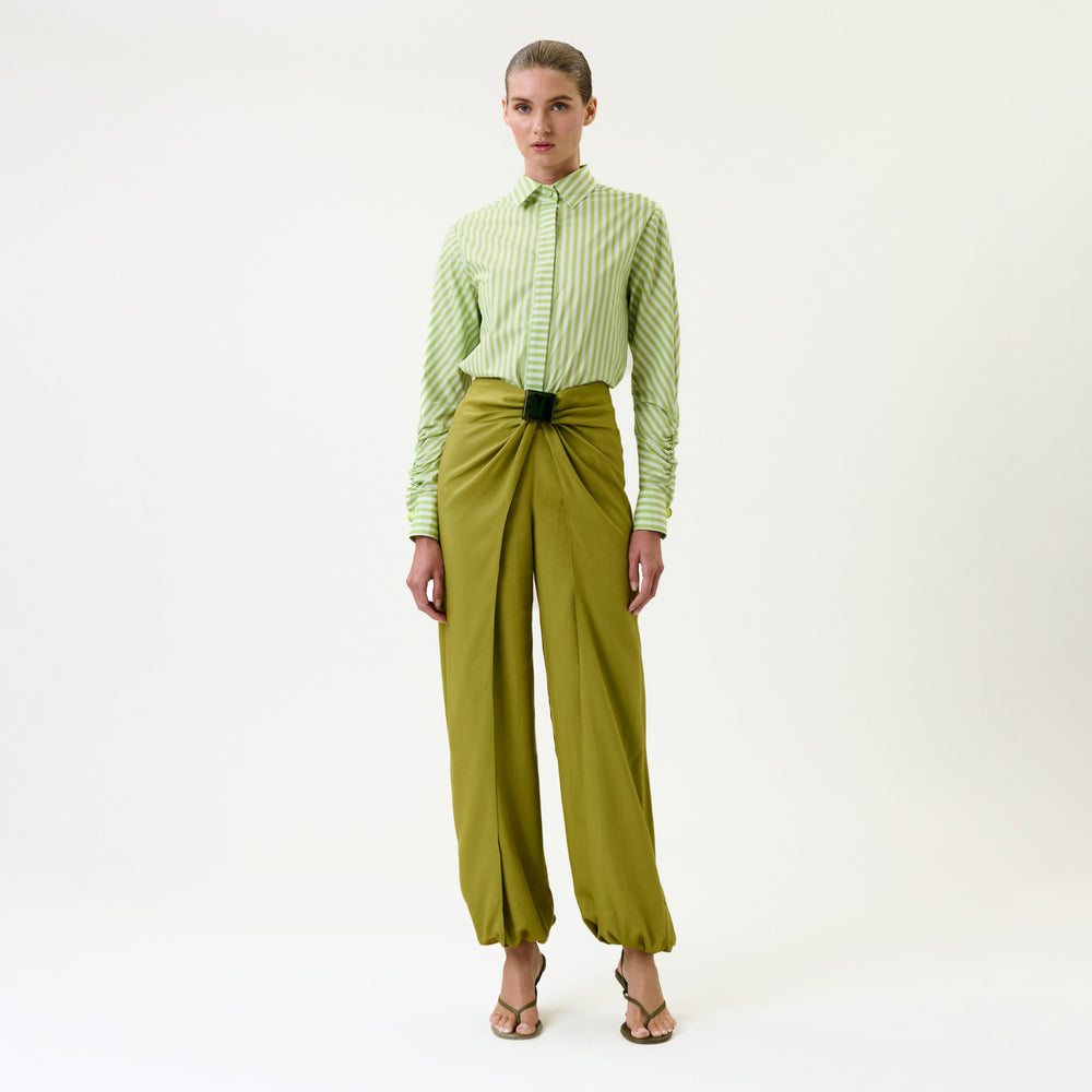 A person stands against a plain background, wearing a green striped shirt tucked into the high-waisted Raimondo Pant in olive green with pleats, gathered ankles, and a dark green belt, paired with sandals.