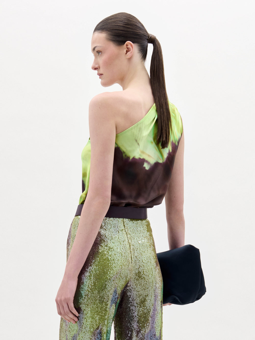 A woman stands against a plain background wearing the Yanet Blouse, a one-shoulder green and brown top, paired with sequined pants, a wide belt with a large buckle, and holding a black clutch.