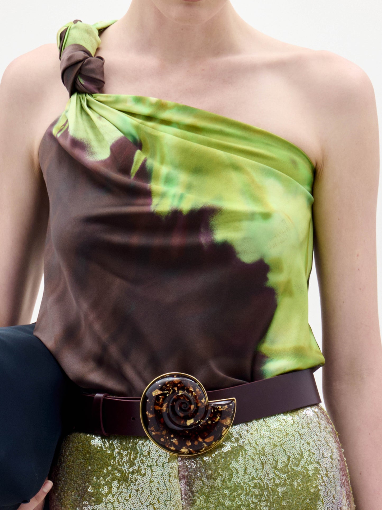 A woman stands against a plain background wearing the Yanet Blouse, a one-shoulder green and brown top, paired with sequined pants, a wide belt with a large buckle, and holding a black clutch.
