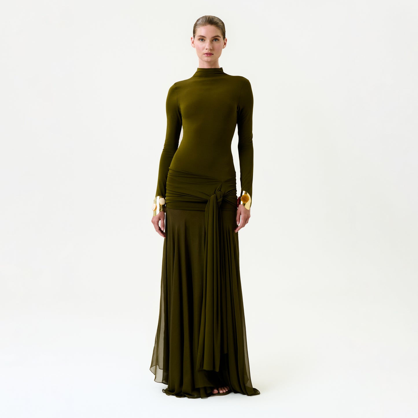 A woman stands against a plain light background, wearing the Monie Dress—an olive green maxi with long sleeves, a high neckline, gold-accented cuffs, Italian fabric, and draped gathers at the hip.