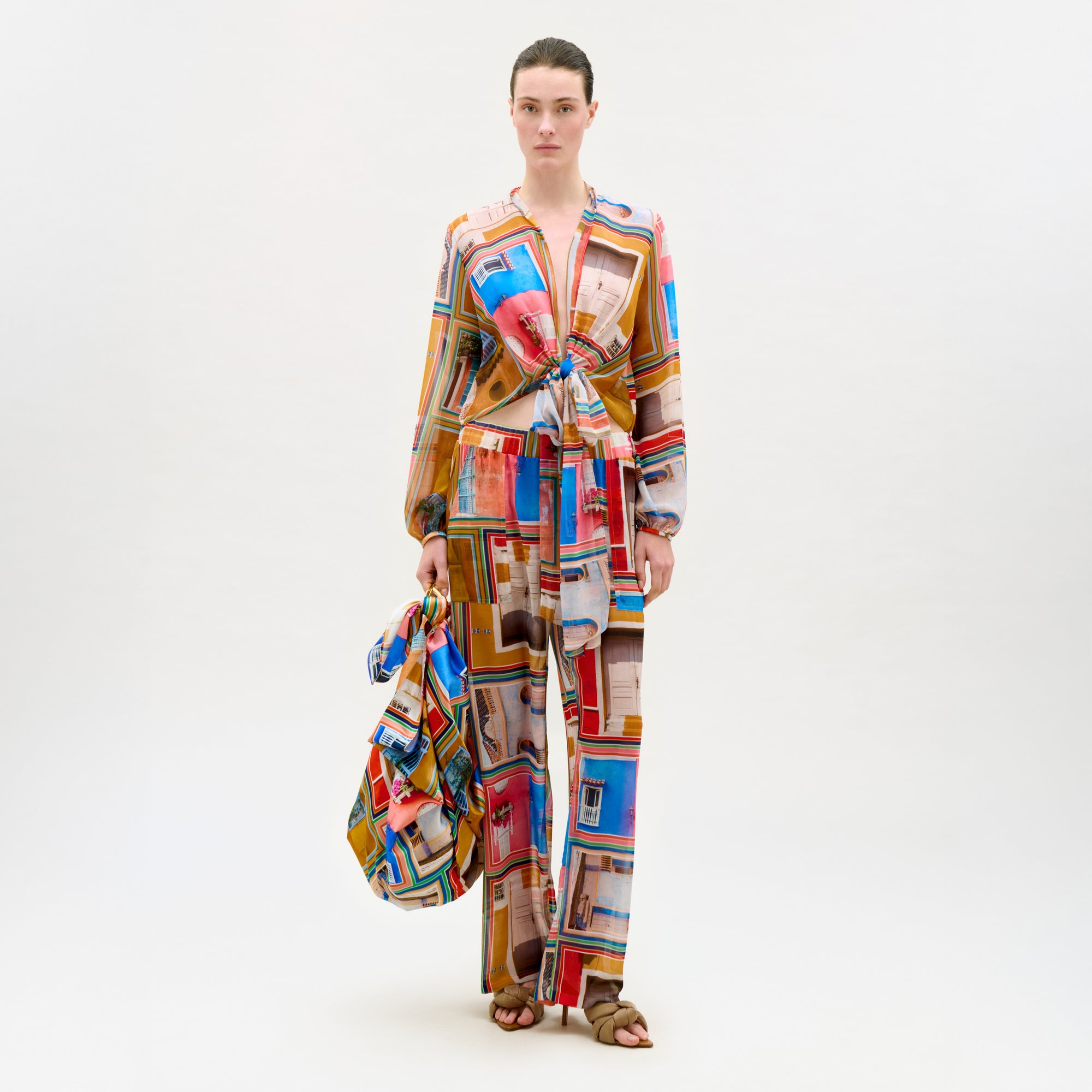 A woman stands against a white background wearing the Nerea Pant, a colorful Italian fabric outfit with window patterns, holding a matching bag. She has brown sandals and her hair is pulled back.
