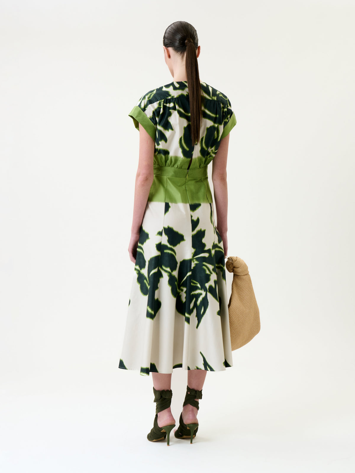 A woman with long dark hair in a ponytail is seen from behind, standing against a plain white background, wearing the Maida Dress—a green and white floral dress—paired with green heels and holding a large tan woven bag.