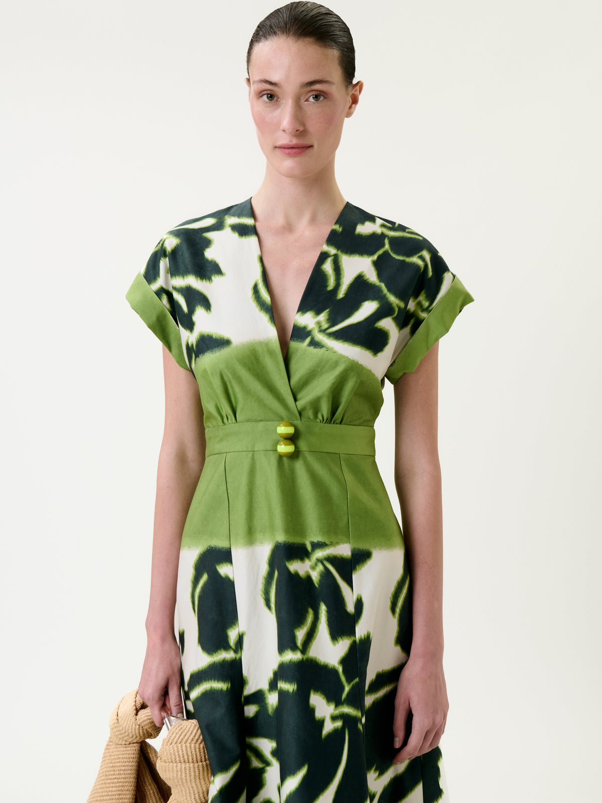 A woman stands against a plain background, holding a textured beige bag in her left hand and wearing the Maida Dress—a green and black patterned dress featuring short sleeves and a V-neckline.