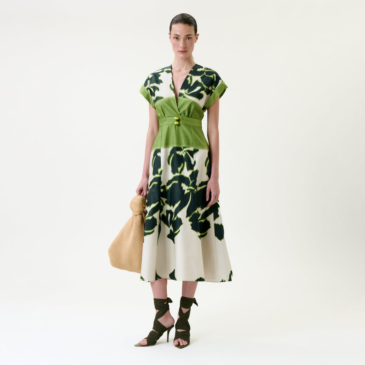 Woman in the Maida Dress—featuring a cream base with green florals—styled with a green belt and green strappy heels, holding a woven tan handbag, standing against a plain white background.