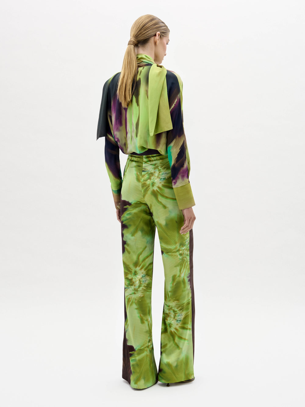 A woman stands against a plain background wearing the Andie Pant, featuring a green and brown abstract print, paired with a matching blouse, wide belt, and scarf.