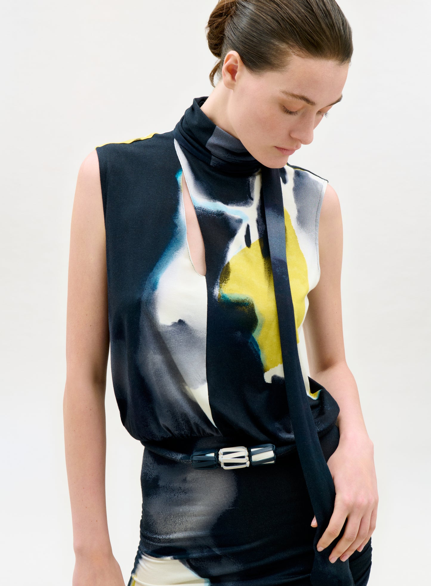 Woman wearing a sleeveless top with abstract design and black scarf against a white background