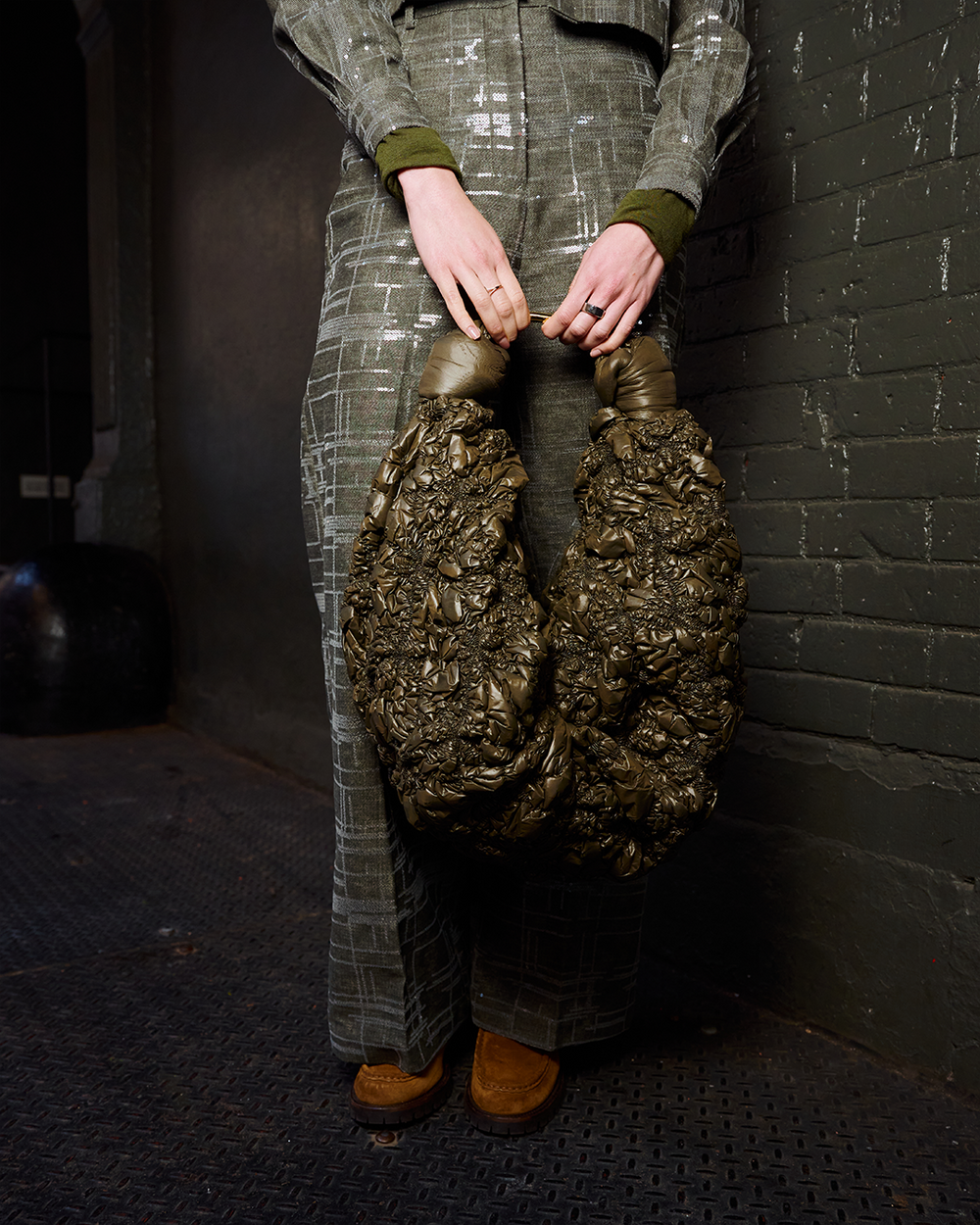 A person wearing a textured gray plaid jumpsuit holds a large, sculptural metallic bronze bag with two bulbous handles, standing against a dark brick wall.