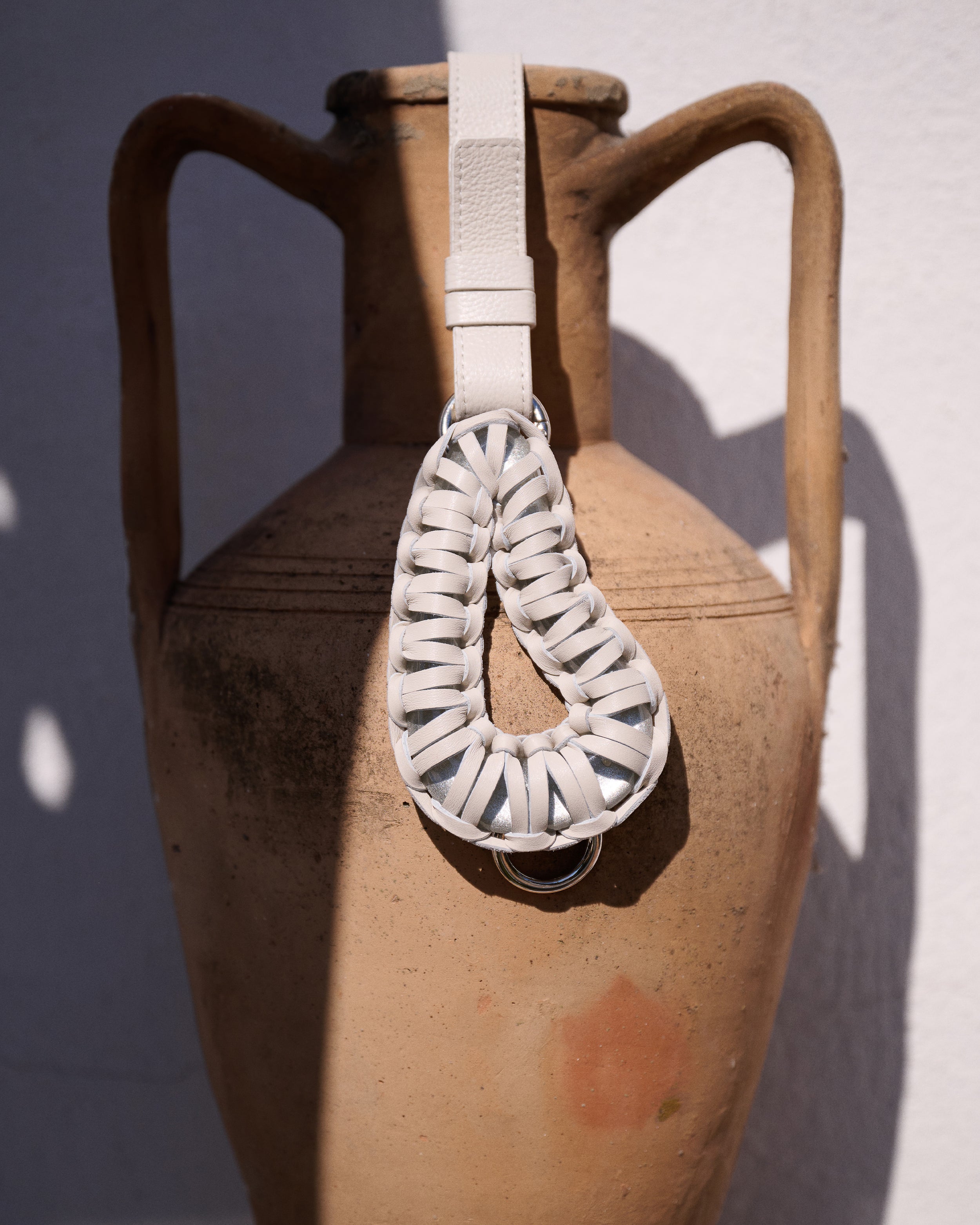 A white woven leather shoulder bag with a single strap, draped over a large rustic terracotta amphora vase against a white wall.