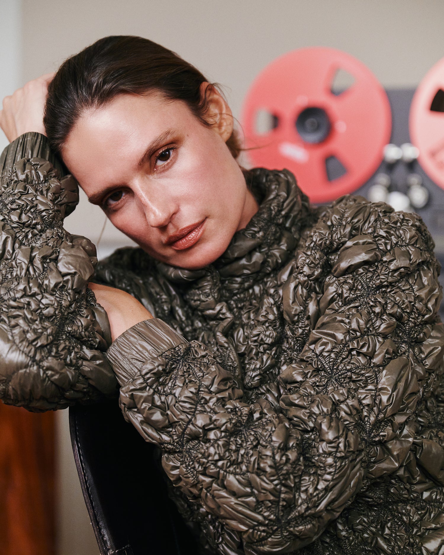 A close-up lifestyle shot of a model wearing a dark brown, heavily textured crinkled quilted jacket. The background features a warm, neutral wall with decorative red film reels.