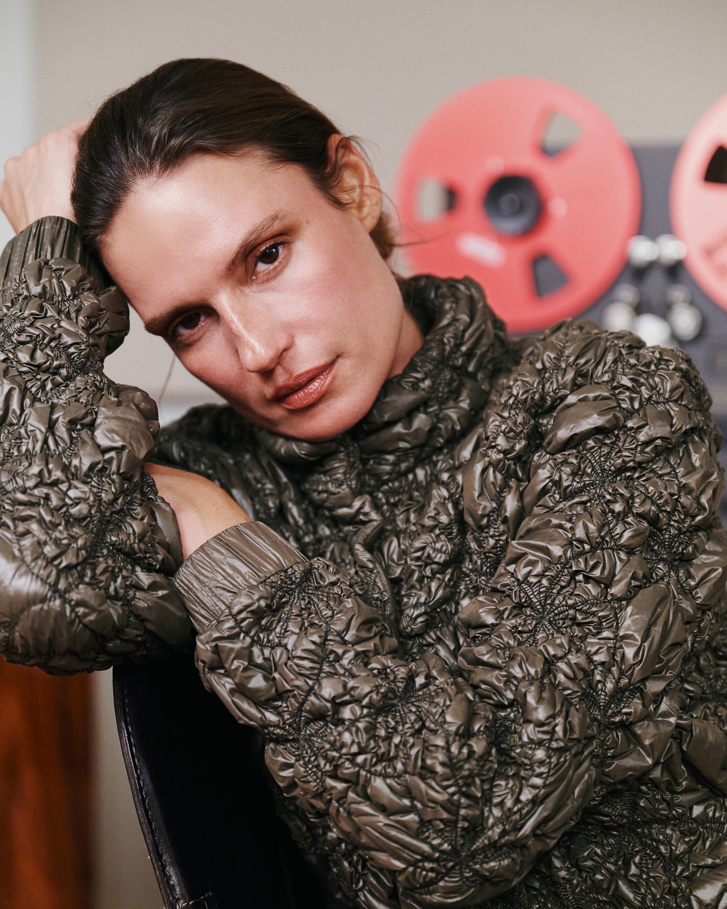 A close-up lifestyle shot of a model wearing a dark brown, heavily textured crinkled quilted jacket. The background features a warm, neutral wall with decorative red film reels.