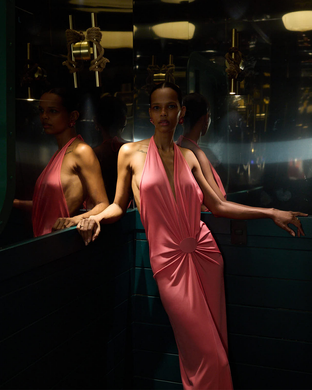 A model wearing a draped, halter-neck pink gown with a gathered waist detail, posing in front of dark mirrored walls that reflect her figure.