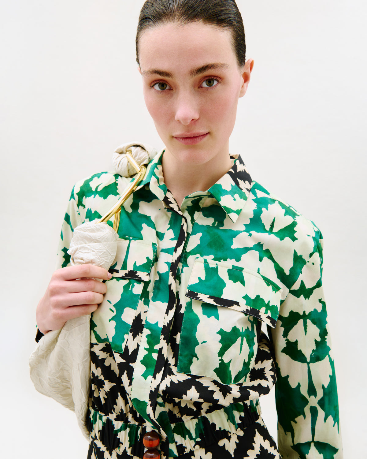 A model wearing a green, black, and cream abstract patterned shirt, holding a textured cream fabric bag against a plain white studio background.