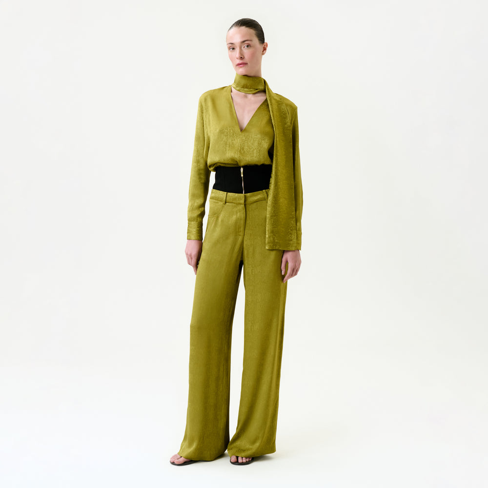 A woman stands against a plain white background wearing the Karel Pant—olive-green, wide-leg trousers in Italian fabric—paired with a matching long-sleeve blouse, scarf, and black belt. The outfit is true to size. Her hair is pulled back and she wears open-toe shoes.