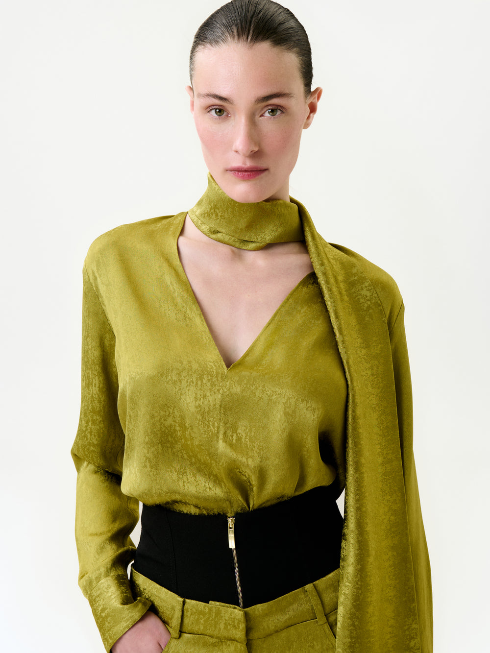 A woman with fair skin and dark slicked-back hair models the Omara Blouse—a shiny olive-green top made from Italian fabric—styled with a matching scarf and high-waisted black pants against a white background.