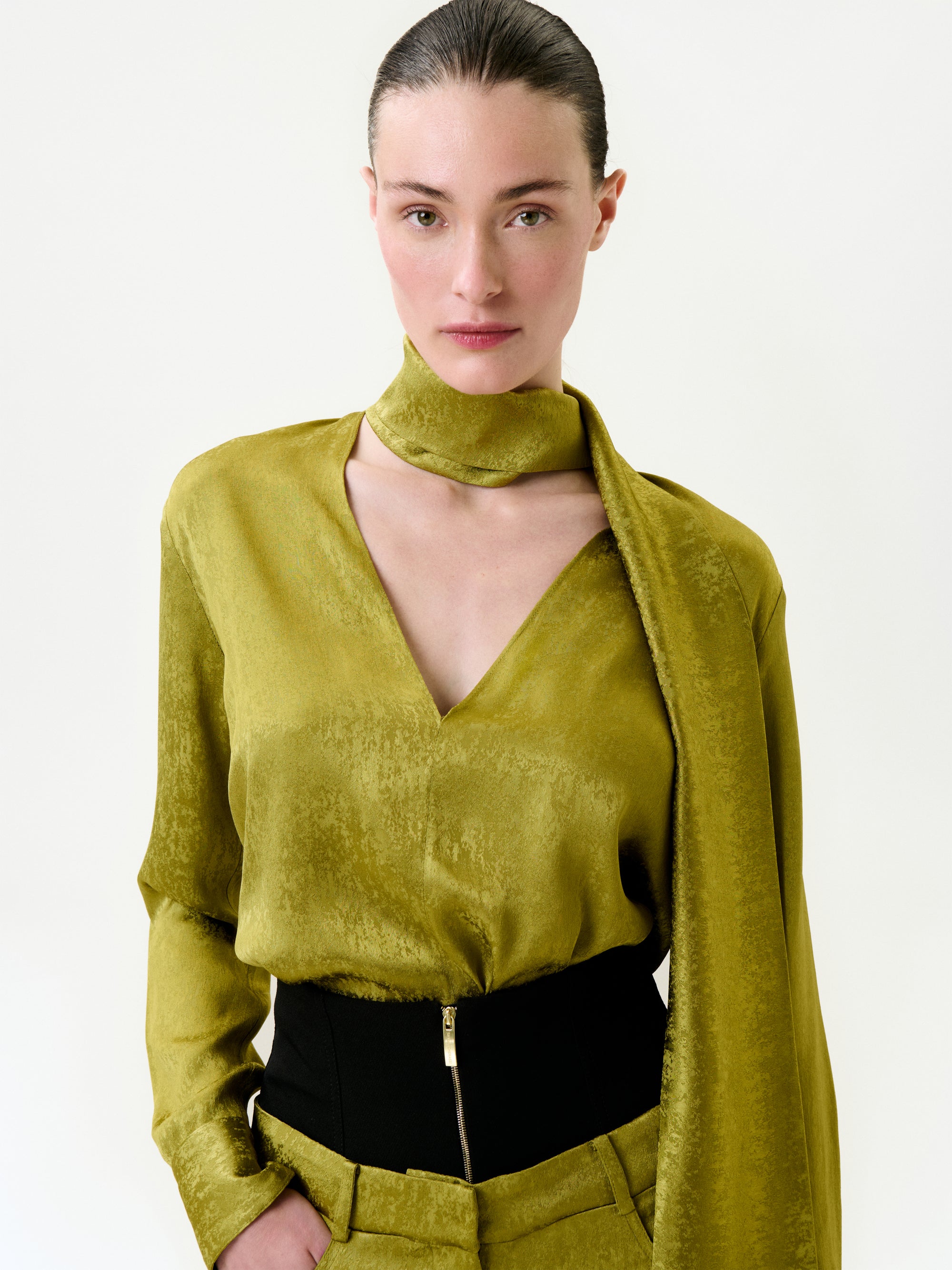 A woman with fair skin and dark slicked-back hair models the Omara Blouse—a shiny olive-green top made from Italian fabric—styled with a matching scarf and high-waisted black pants against a white background.