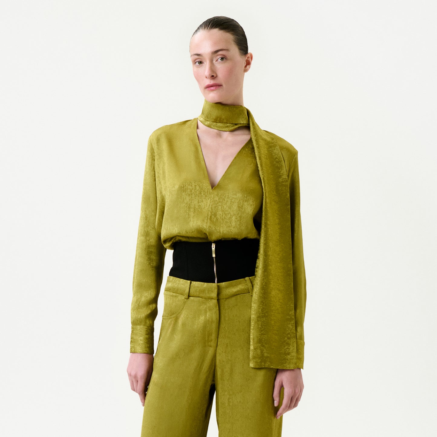 A person with slicked-back hair wears the Omara Blouse—gold-green, textured Italian fabric, deep V-neck—paired with a matching scarf and high-waisted pants featuring a black belt and gold zipper, set against a plain white background.