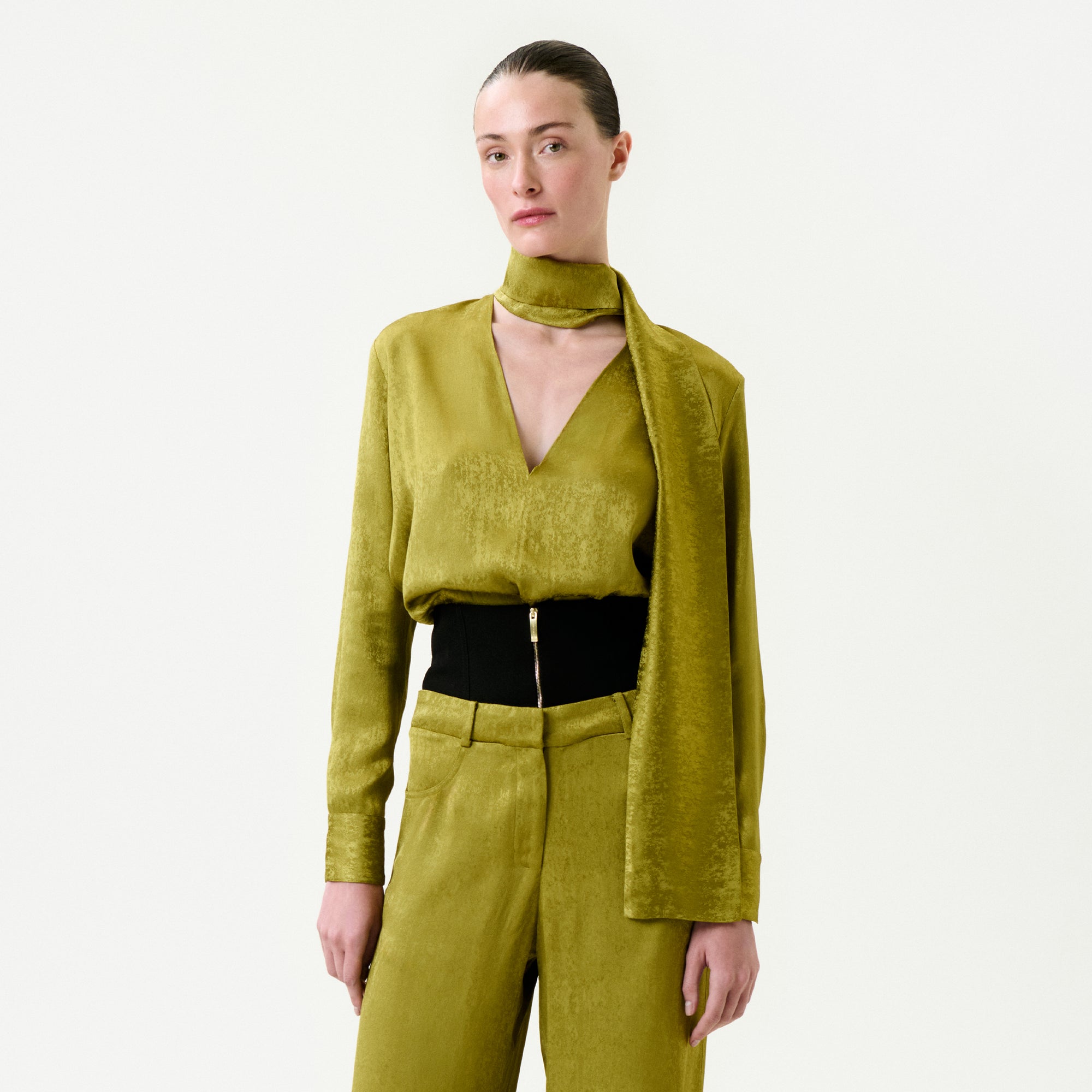 A person with slicked-back hair wears the Omara Blouse—gold-green, textured Italian fabric, deep V-neck—paired with a matching scarf and high-waisted pants featuring a black belt and gold zipper, set against a plain white background.