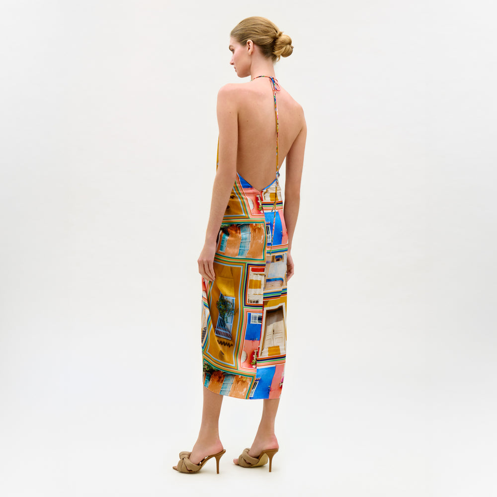 A woman with blonde hair in a neat bun wears the colorful silk Guadalupe Dress with a halter neckline and beige high-heeled sandals, standing against a plain white background.