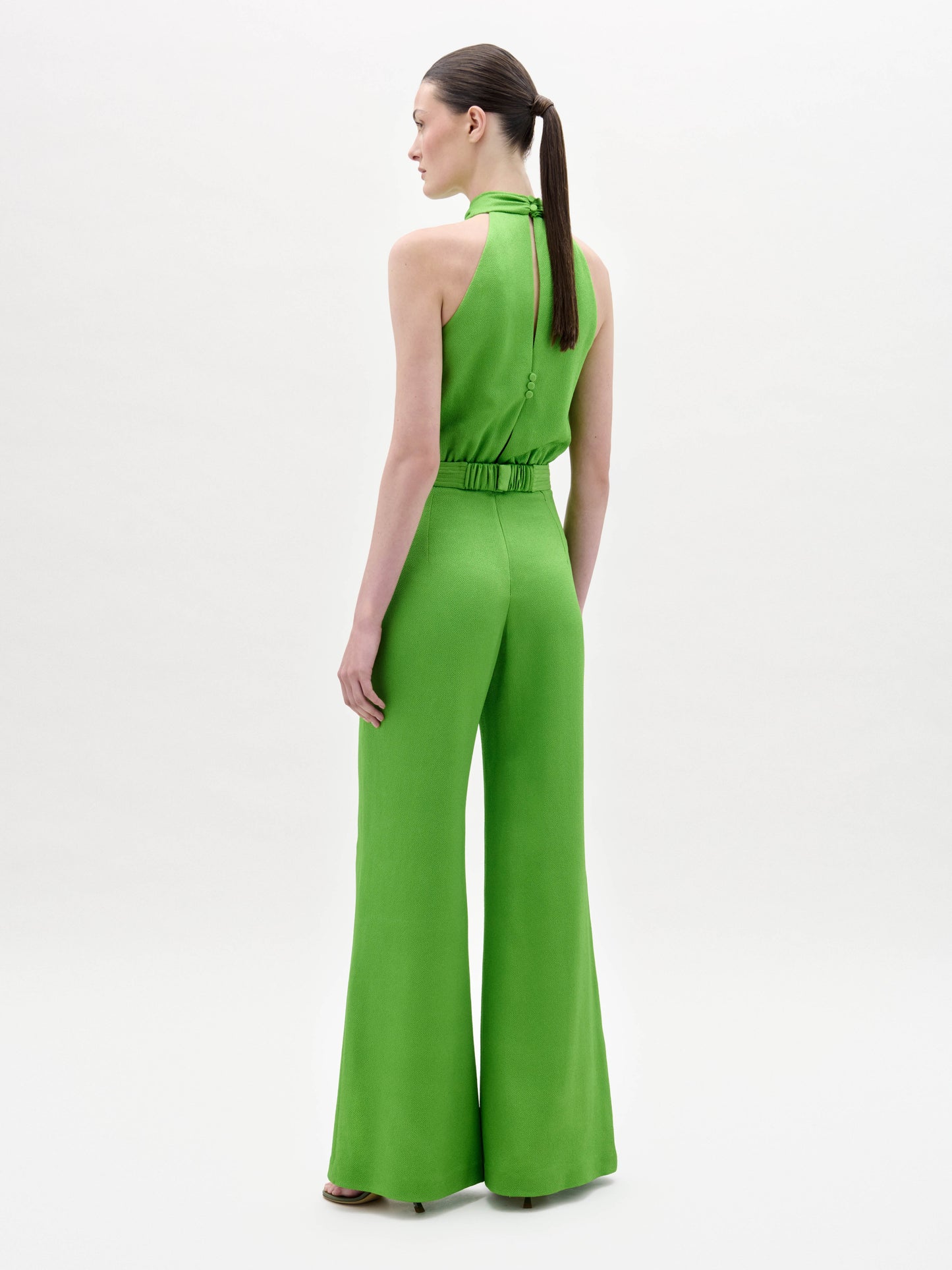 A woman stands facing forward in the Siv Jumpsuit, a bright green halter-neck style with wide-leg pants and a matching belt, paired with black sandals, against a plain white background.