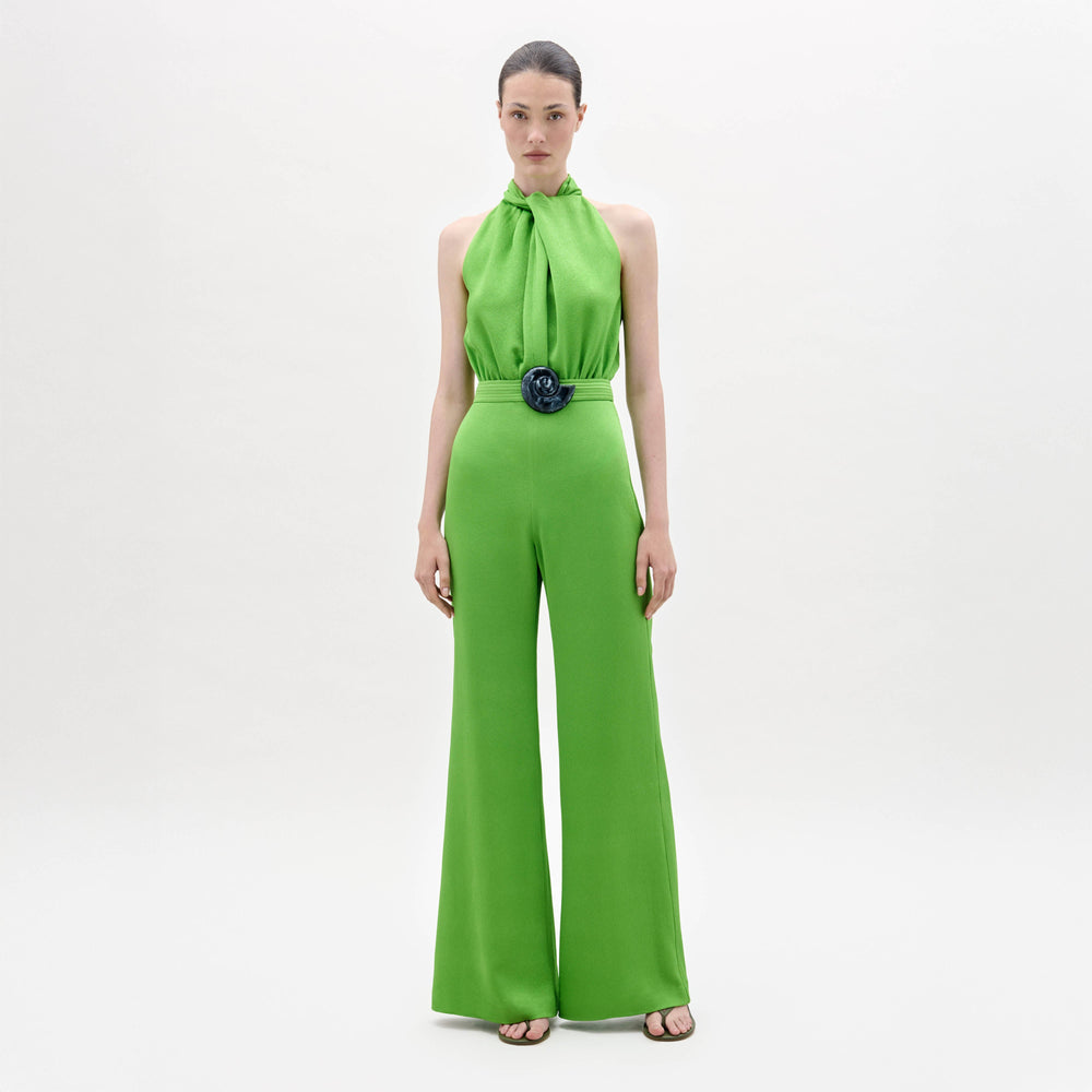 A woman stands facing forward in the Siv Jumpsuit, a bright green halter-neck style with wide-leg pants and a matching belt, paired with black sandals, against a plain white background.