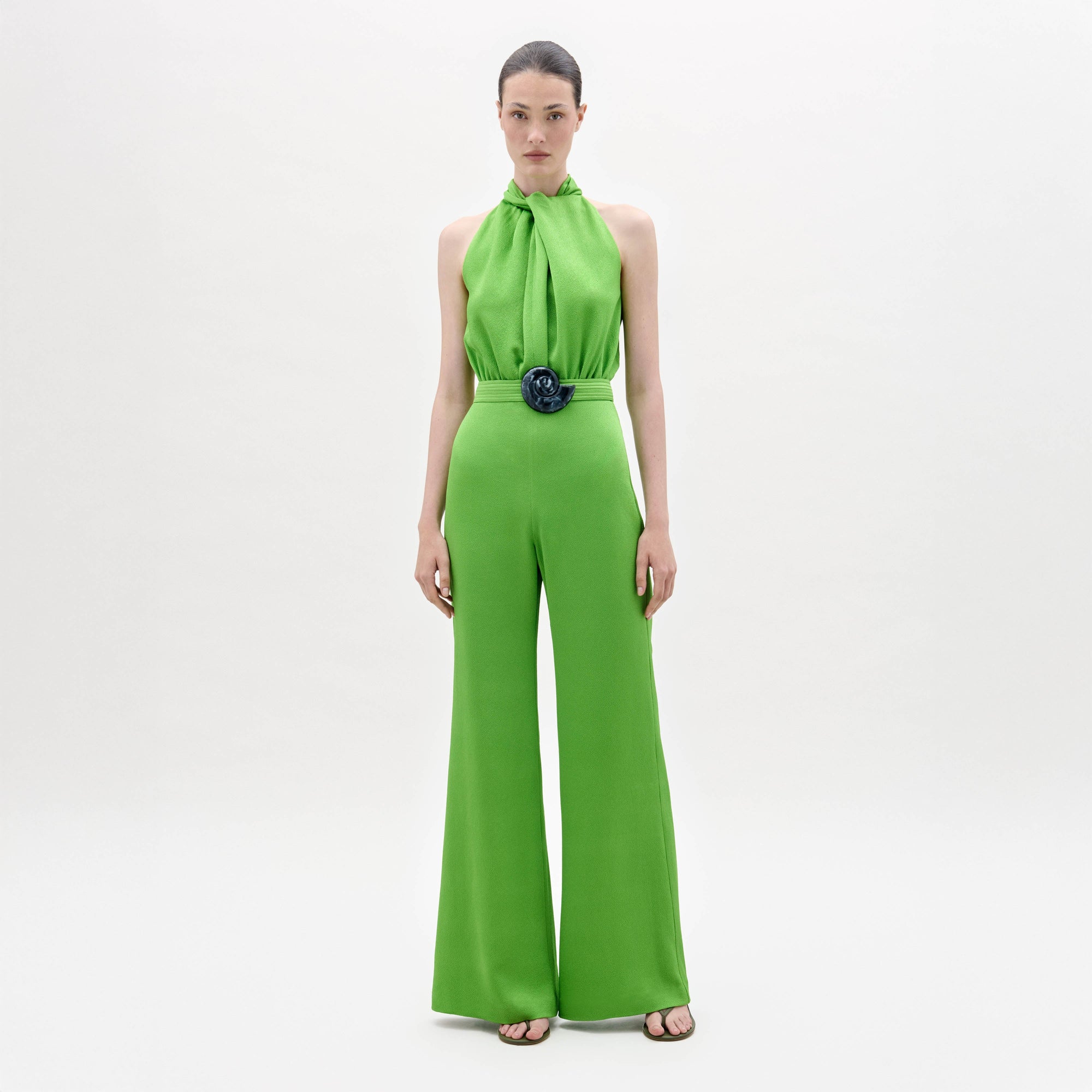 A woman stands facing forward in the Siv Jumpsuit, a bright green halter-neck style with wide-leg pants and a matching belt, paired with black sandals, against a plain white background.