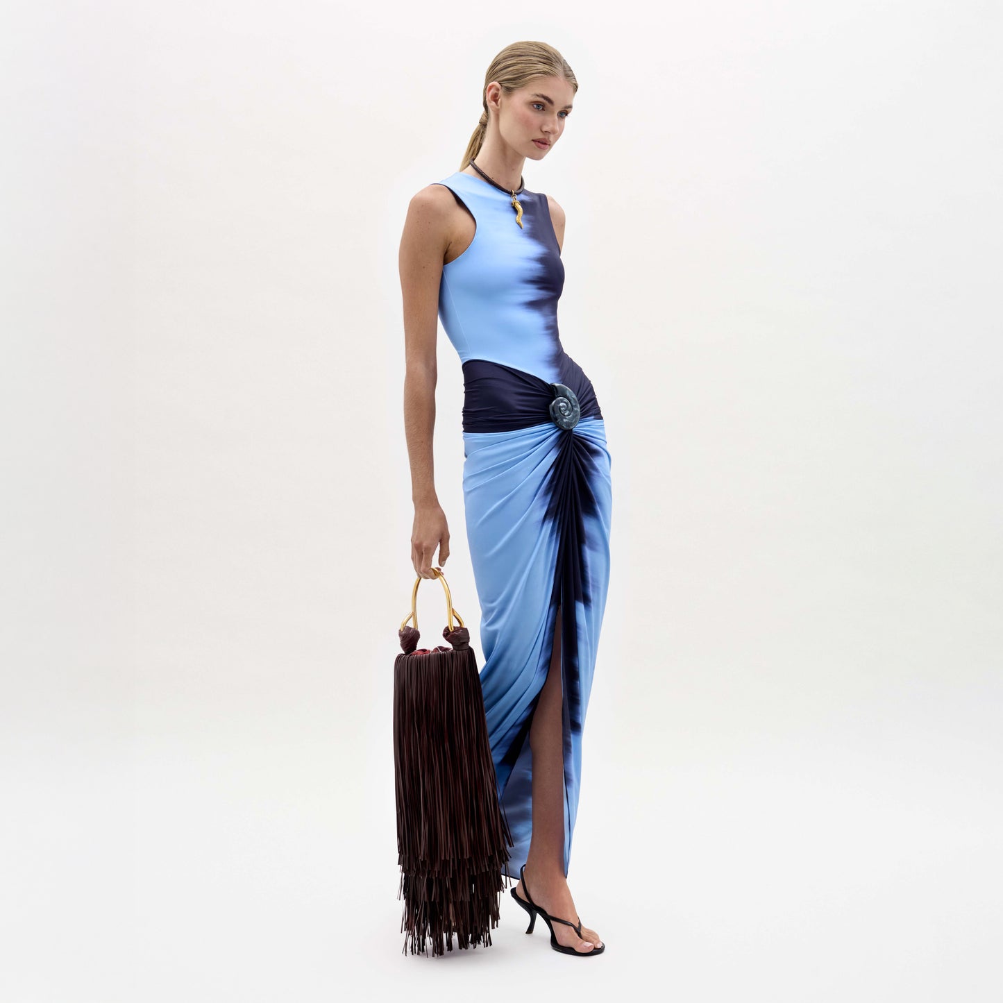 A woman wears the Leni Dress, a blue gradient midi with a high slit, and carries a large brown fringed bag. Crafted from luxurious Italian fabric, she completes the look with black strappy heels against a plain white background.