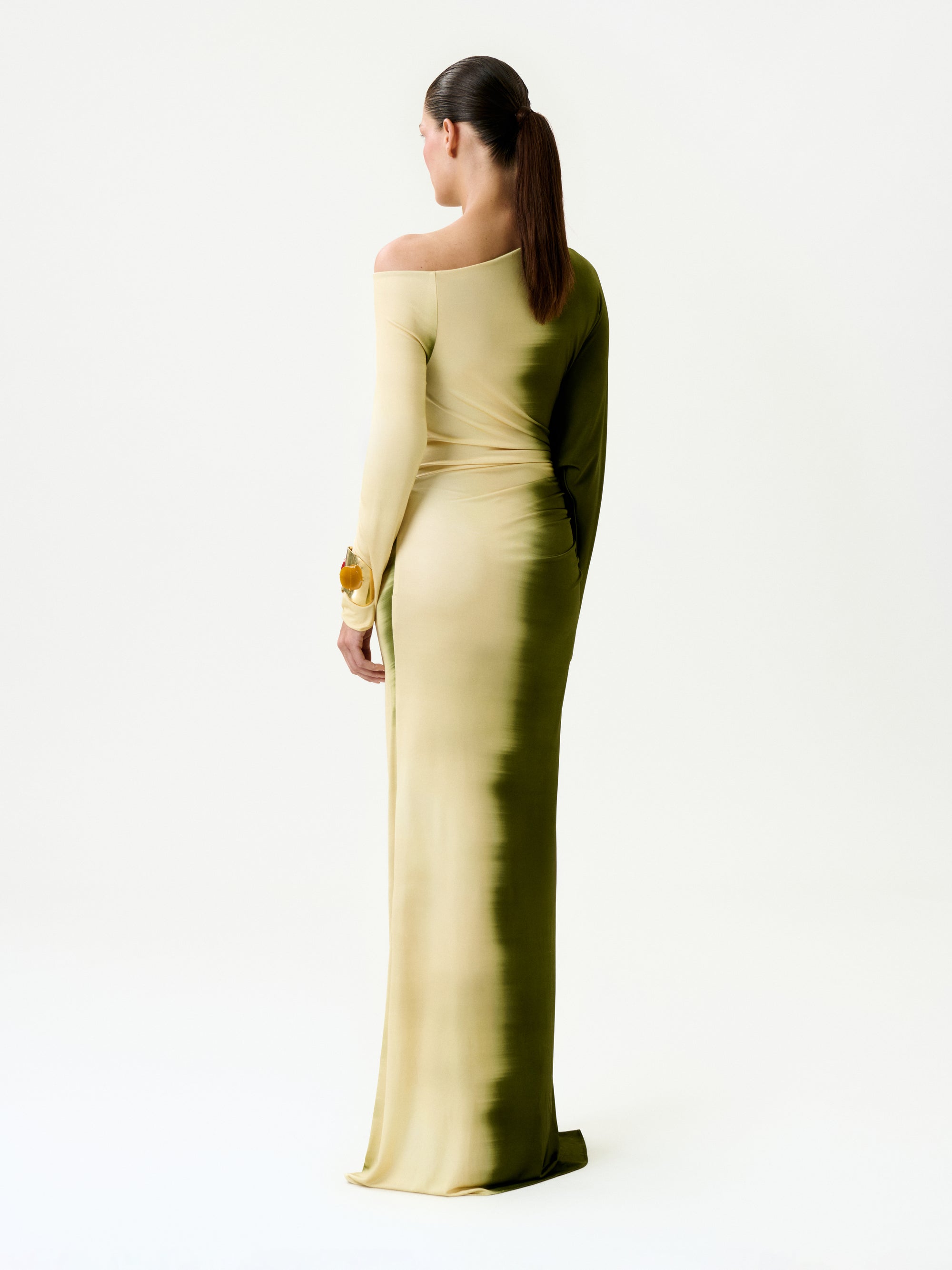 A person with long brown hair in a ponytail stands with their back to the camera against a white background, wearing the Nadja Dress—a form-fitting, off-shoulder ombré yellow and green maxi dress made in Colombia.