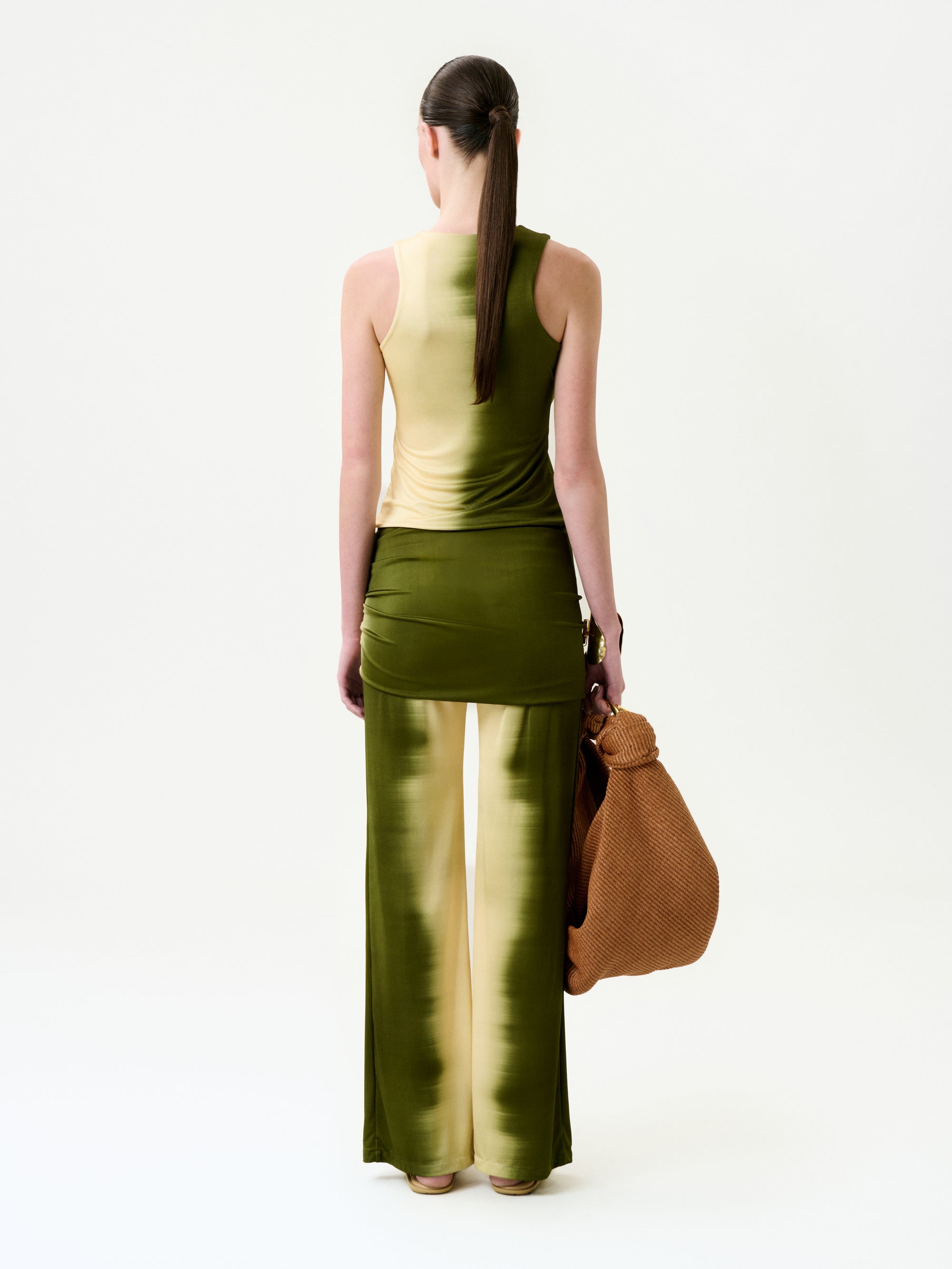 A woman with a long ponytail faces away, wearing the Sonu Pant—wide-leg, sleeveless yellow-green gradient pants made in Colombia—with a matching top. She holds a large brown woven bag in her left hand against a plain white background.