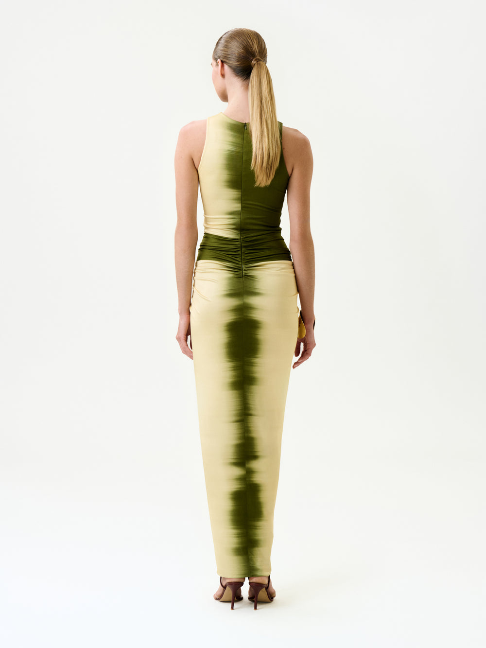 A woman with straight blonde hair in a low ponytail stands facing away, wearing the Leni Dress—a sleeveless midi dress made from Italian fabric featuring a vertical green and cream tie-dye pattern—in a minimal white studio.
