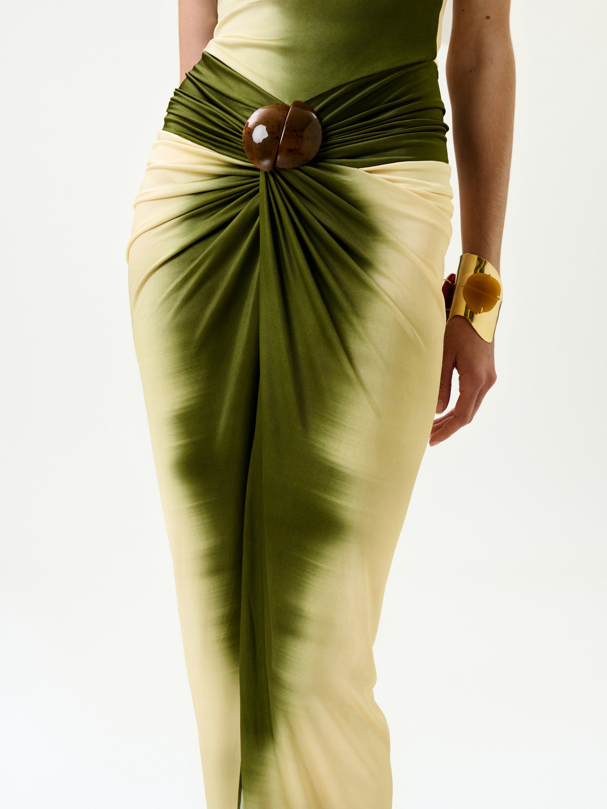 A person wearing the Leni Dress, a green and cream draped skirt made from Italian fabric with a large round brown buckle at the waist, paired with a chunky yellow bracelet, stands against a plain white background.
