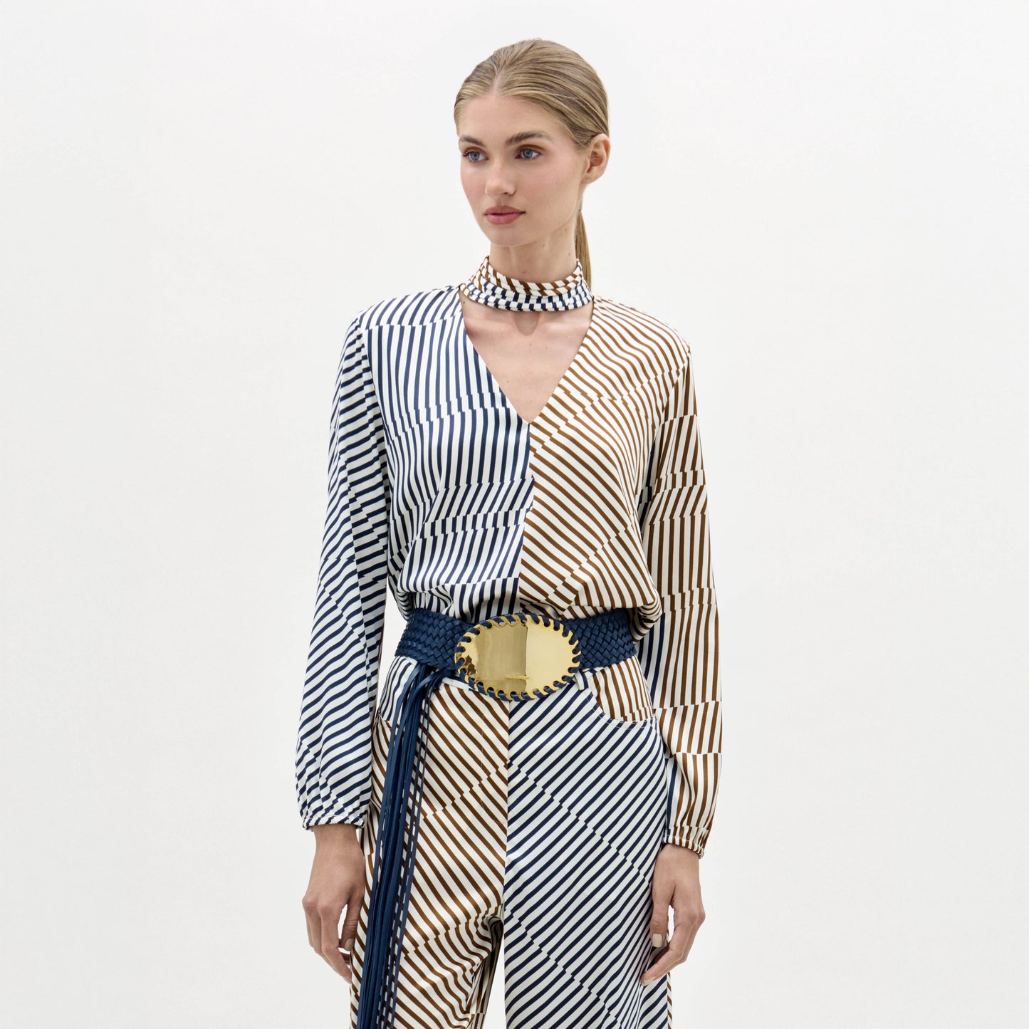 Woman wearing the Jasper Blouse, a long-sleeved, blue and beige striped top, paired with a matching choker and a wide belt with a large gold buckle, stands against a white background.