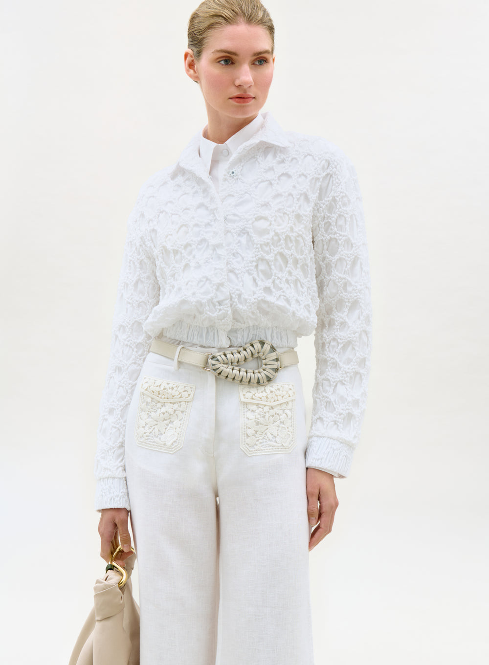 Woman wearing a white lace top and pants with a beige handbag on a light background