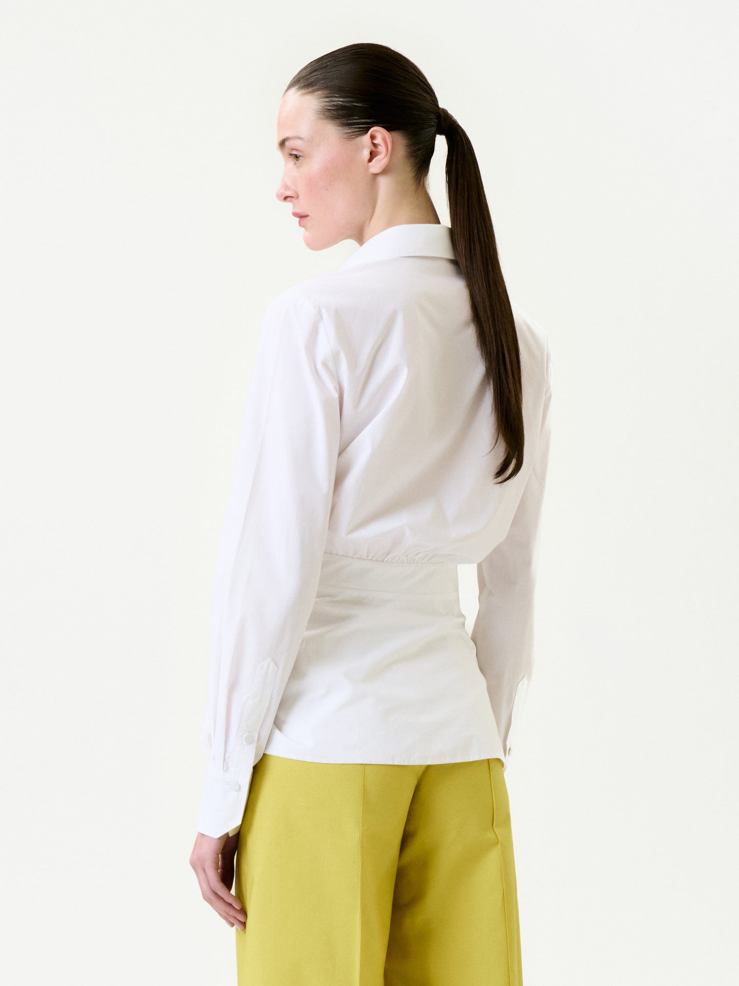 A woman with straight brown hair in a ponytail, seen from behind, wears the Malen Shirt—a white 100% cotton collared shirt—paired with yellow trousers against a plain light background.