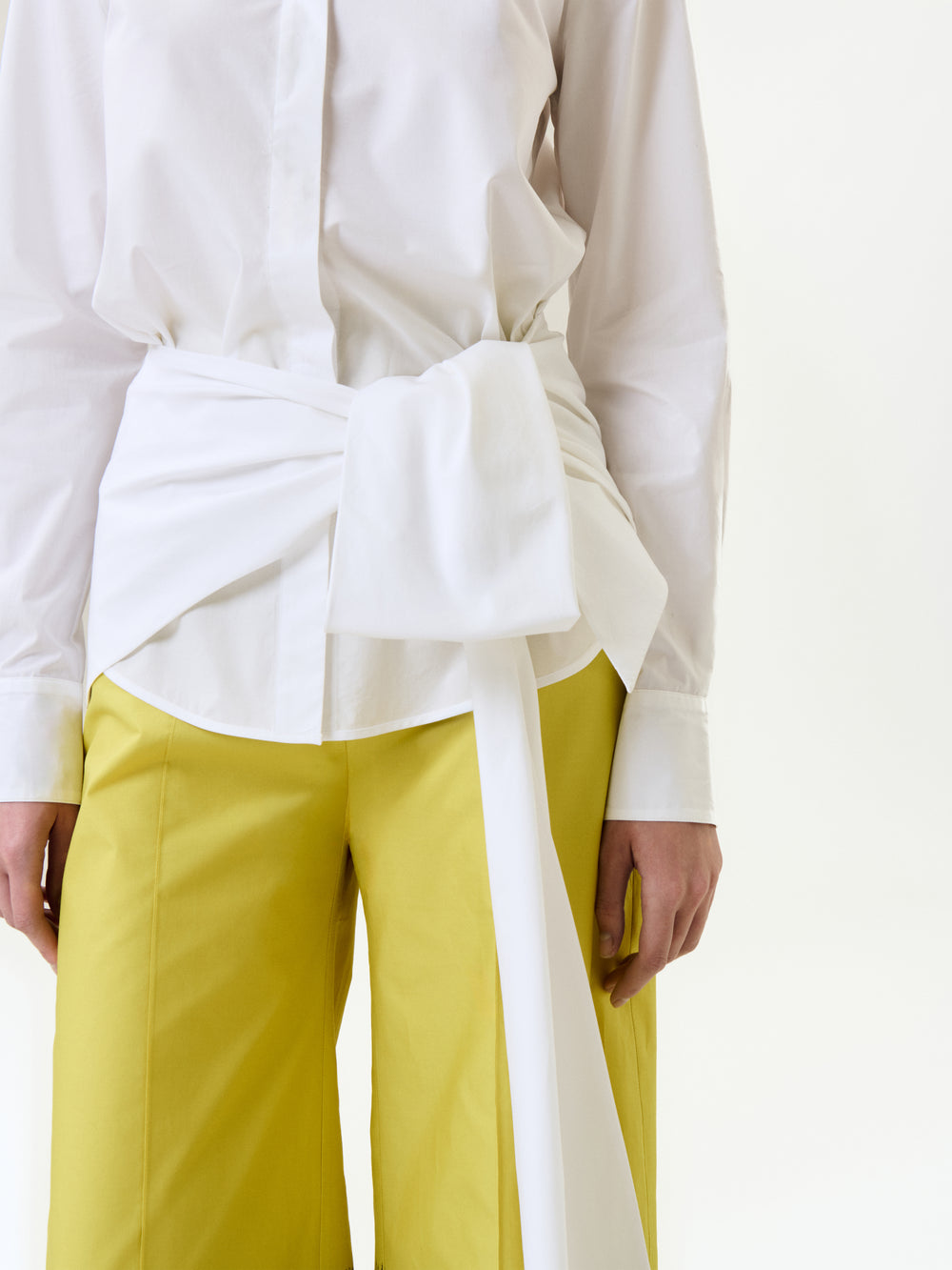 A person wears the Malen Shirt—a white button-up made from Portuguese fabric with a wide waist belt—paired with bright yellow pants. Only the torso and upper legs are shown against a plain white background.