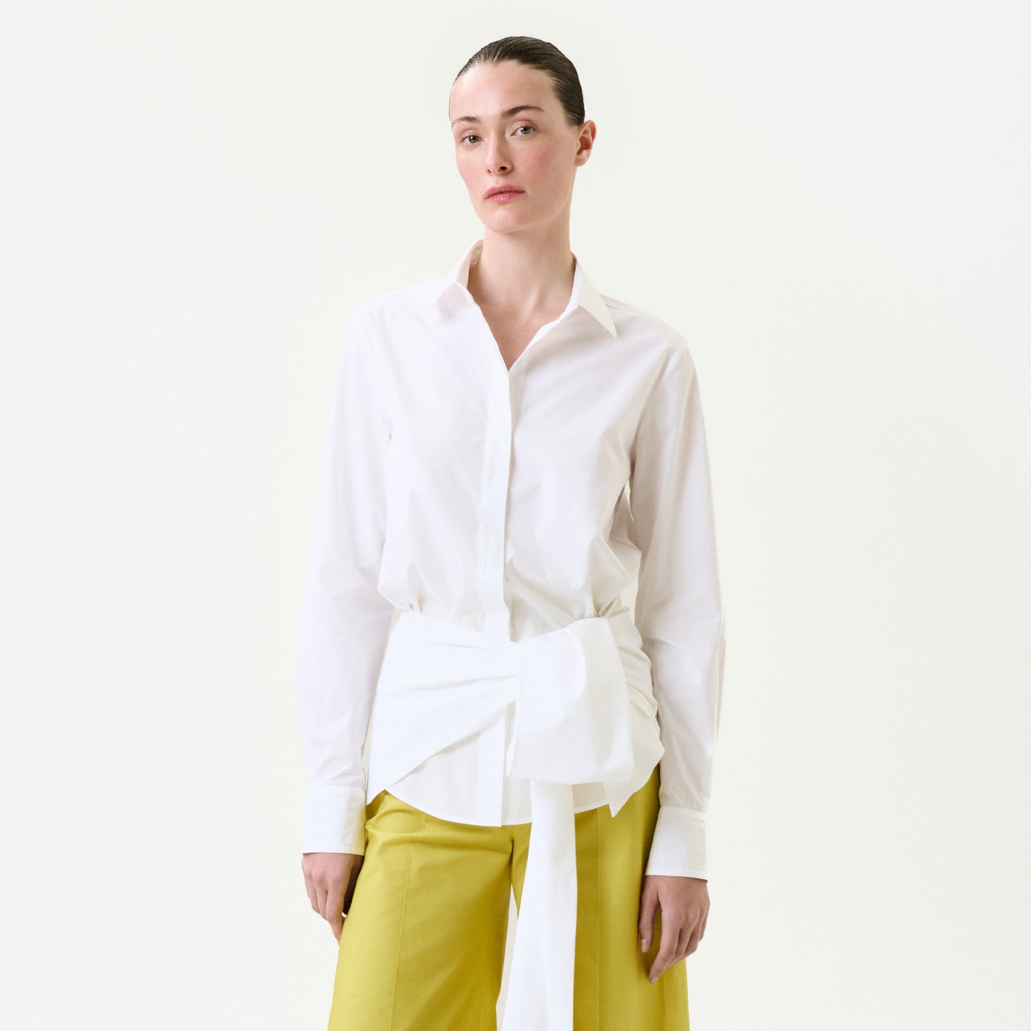 A person with slicked-back hair and a neutral expression stands against a plain white background, wearing the Malen Shirt—a white 100% cotton button-up with a large tied sash at the waist—paired with yellow wide-leg pants.